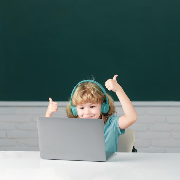 Un chico inteligente con los pulgares hacia arriba en los auriculares se sienta en el escritorio para estudiar en línea en una computadora portátil en la escuela.