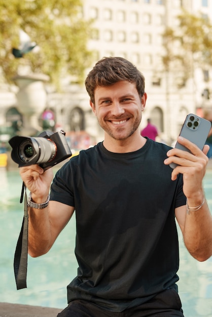 Chico joven alegre sentado cerca de la fuente con cámara de fotos moderna y smartphone