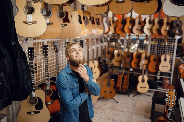 Chico joven barbudo que elige la guitarra acústica en la tienda de música.
