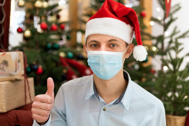 Un chico joven con una camisa y un sombrero de santa claus en el fondo de un árbol de Navidad. Vacaciones durante una pandemia