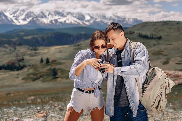 Un chico joven con una chaqueta de mezclilla con una mochila se para junto a una chica con pantalones cortos y una camisa y muestra sus fotos de la cámara. Fondo de montañas y valle verde.