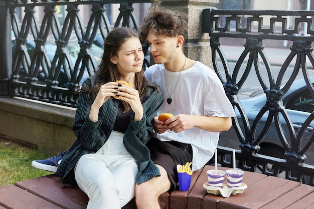 Un chico joven y una chica de la ciudad están sentados en un banco y comen hamburguesas.