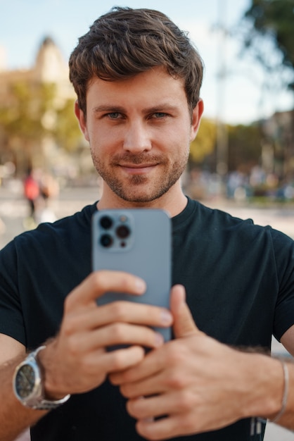 Chico joven confidente que toma la foto en el teléfono inteligente en el parque de la ciudad