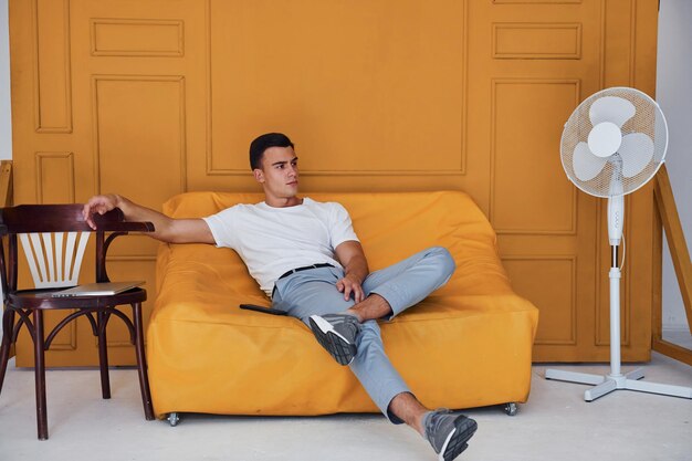 Chico joven con corte de pelo elegante y ropa informal sentado en el interior de la habitación con interior de color naranja