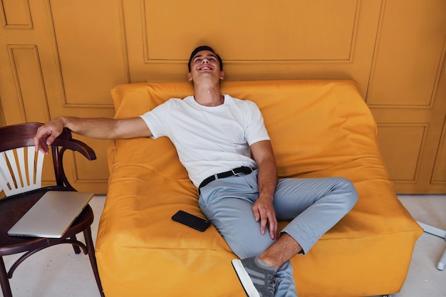 Chico joven con corte de pelo elegante y ropa informal sentado en el interior de la habitación con interior de color naranja