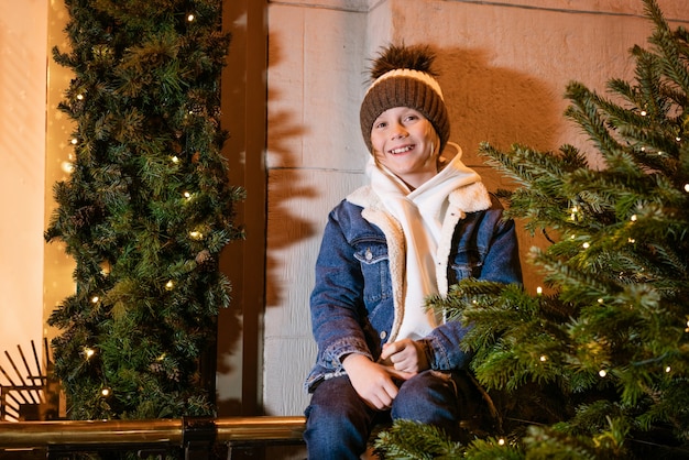 Chico joven en elegante chaqueta con sombrero de moda se encuentra en la ciudad cerca del edificio decorado con christm ...