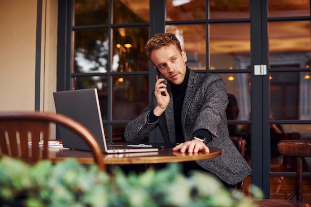 Chico joven elegante en ropa formal se sienta en la cafetería con su computadora portátil y habla por teléfono.