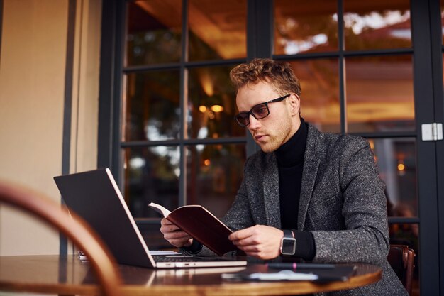 Chico joven elegante en ropa formal se sienta en la cafetería con su computadora portátil y lee el libro.