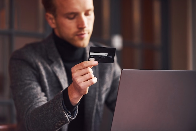 Chico joven elegante en ropa formal se sienta en la cafetería con su computadora portátil y tarjeta de crédito en la mano.