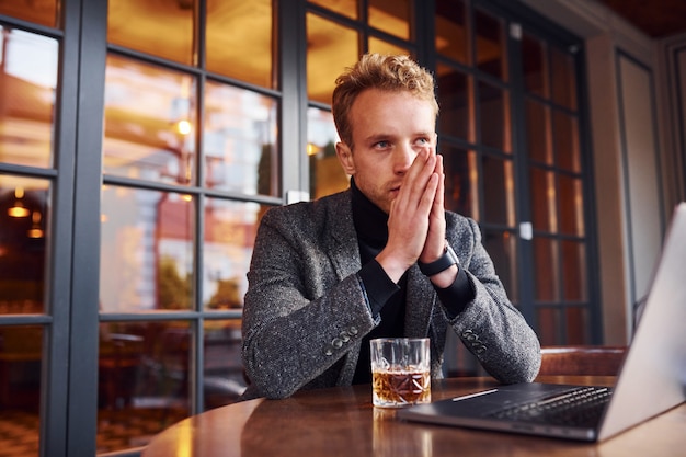 Chico joven elegante en ropa formal se sienta en la cafetería con su computadora portátil y vaso de alcohol.