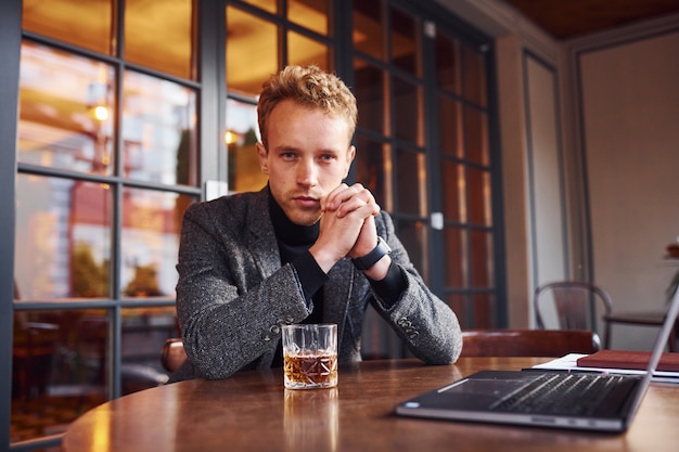 Chico joven elegante en ropa formal se sienta en la cafetería con su computadora portátil y vaso de alcohol.