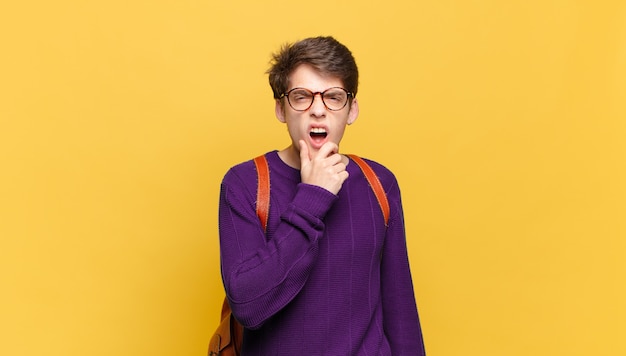 Chico joven estudiante con la boca y los ojos bien abiertos y la mano en la barbilla sintiéndose desagradablemente sorprendido diciendo qué o guau