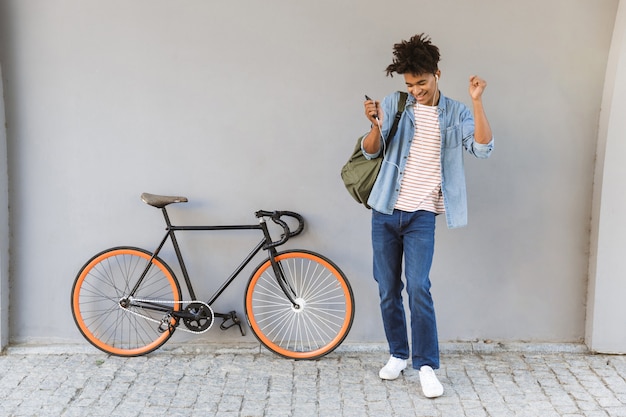 Chico joven feliz caminando al aire libre con bicicleta escuchando música charlando por teléfono móvil