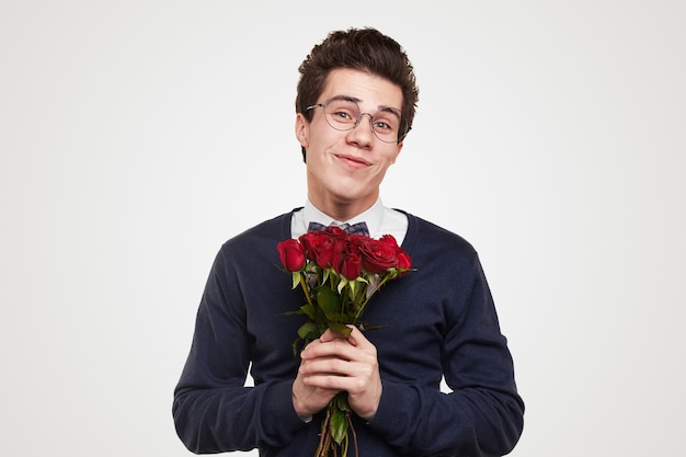 Chico joven feliz en ropa elegante y gafas con ramo de rosas rojas en las manos sonriendo y mirando