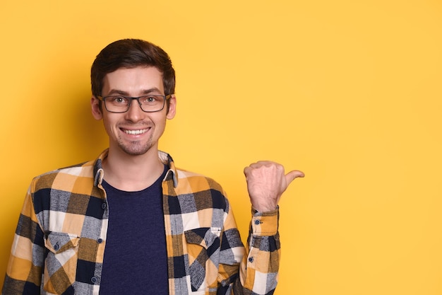 Chico joven en gafas apuntando con el dedo al espacio sobre fondo de estudio amarillo