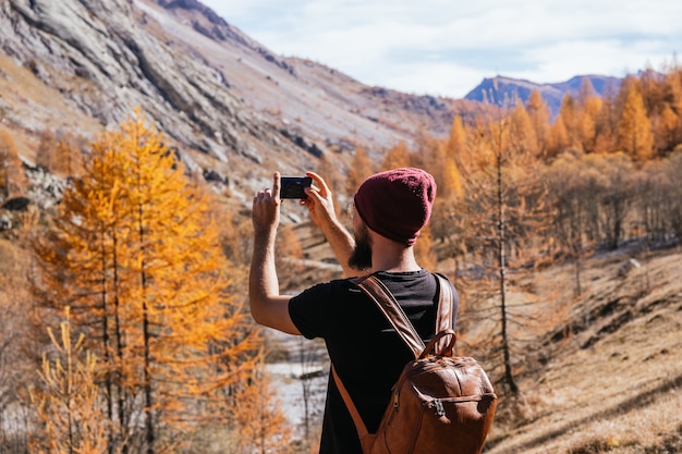 Chico joven con gafas y barba está tomando una foto horizontal con su teléfono mientras camina por la montaña