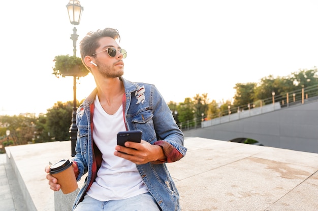 Foto chico joven guapo caminando al aire libre con teléfono móvil, tomando café y escuchando música