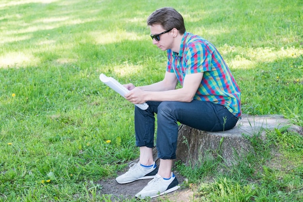 Chico joven guapo con camisa colorida leyendo mientras se sienta en un tocón en un parque verde