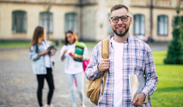 Chico joven guapo estudiante barbudo en ropa casual con mochila y libros en las manos está caminando y posando en los edificios de una universidad