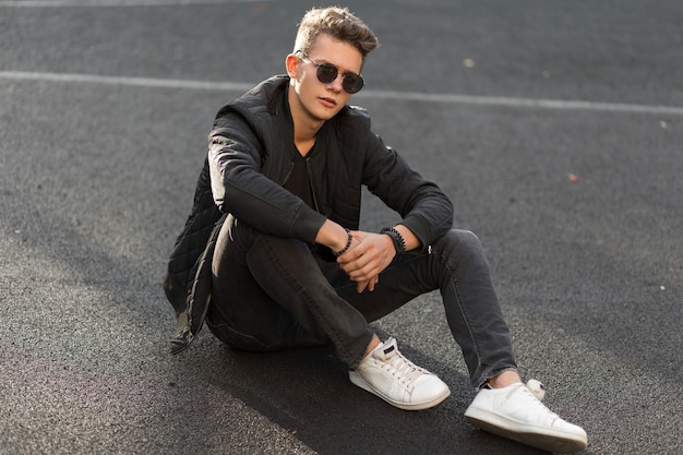 Chico joven guapo hipster con gafas de sol en ropa negra de moda con chaqueta y elegantes zapatillas blancas sentado en la calle