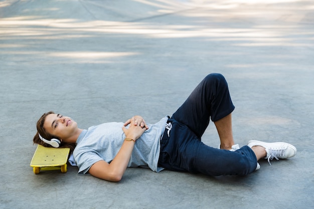 Chico joven guapo pasar tiempo en el parque de patinaje, escuchando música con auriculares, tendido en patineta