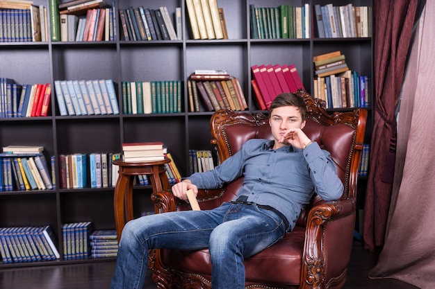 Chico joven guapo en ropa de moda sentado en la silla de la biblioteca mientras sostiene un libro y mira a la cámara