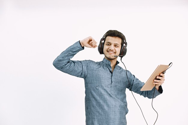 Chico joven Indaian en auriculares escuchando música. Bailando en la pared blanca, aislada.