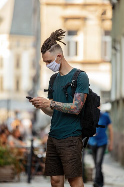 Chico joven con mascarilla y rastas está usando un teléfono móvil en la calle