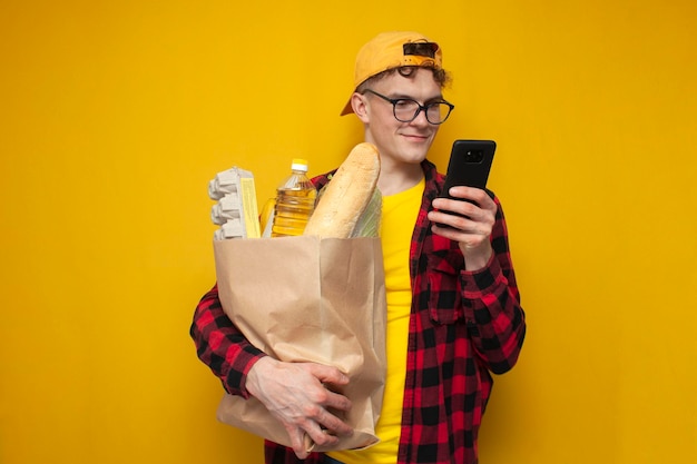 Chico joven con paquete de productos usa teléfono en comprador de fondo amarillo con comida