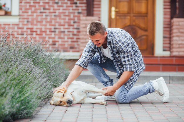 Chico joven con perro perdiguero a pie en el patio de verano. Hombre guapo acariciando a su golden retriever frente a la casa.