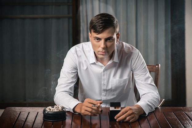 Chico joven posando sentado en una mesa en la que se encuentra un cenicero lleno de cigarrillos