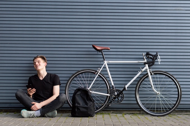 Chico joven sentado con una mochila y un teléfono inteligente cerca de la bicicleta