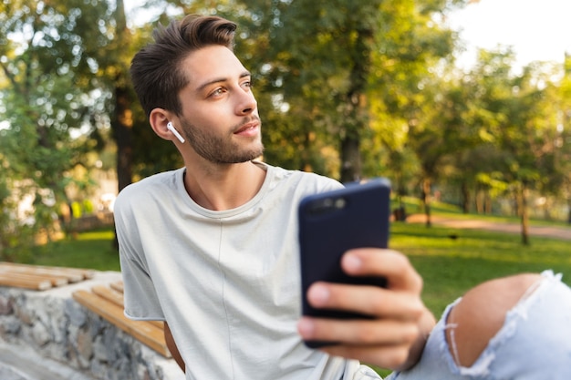 Chico joven sentado en el parque al aire libre con teléfono móvil escuchando música con auriculares