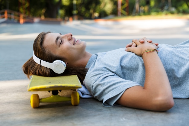 Chico joven sonriente pasar tiempo en el parque de patinaje, escuchando música con auriculares, tendido en patineta