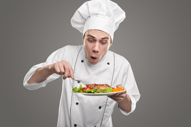 Chico joven sorprendido en uniforme de chef recogiendo tomate del plato mientras saborea un delicioso plato contra el fondo gris