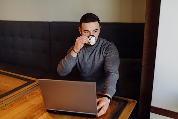 Chico joven con su teléfono móvil y tomando un café. Café expreso de hombre joven de moda en la cafetería de la ciudad durante la hora del almuerzo y trabajando en la computadora portátil