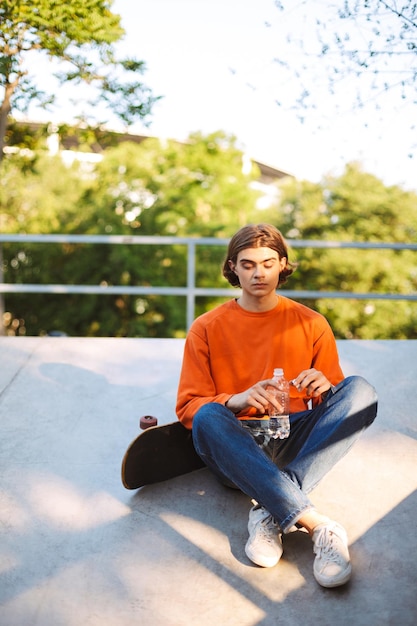 Chico joven en suéter naranja con monopatín sosteniendo una botella de agua en la mano mientras pasa tiempo en el parque de patinaje