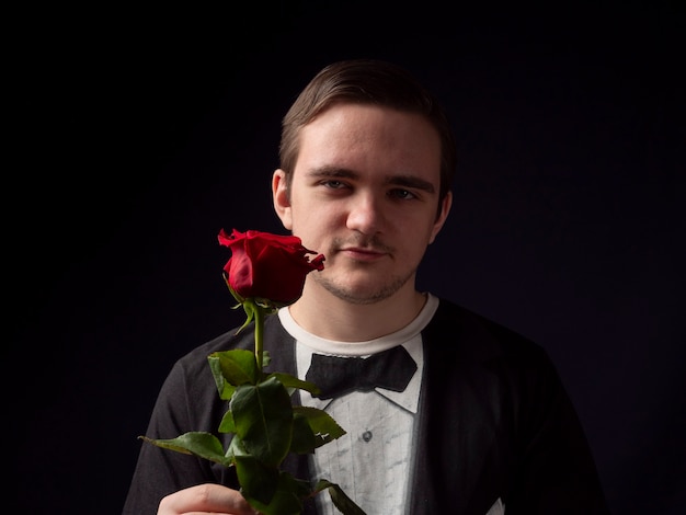 Chico joven con un traje de camiseta negra sostiene una rosa roja en sus manos y sonríe sobre un fondo negro