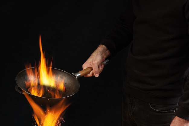 Chico joven vestido con jersey y jeans que sostiene una sartén wok ardiente sobre el fuego contra el estudio negro ...