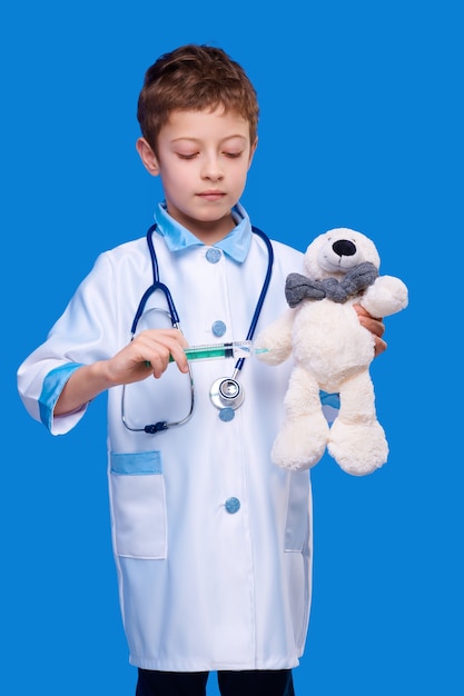Chico lindo en abrigo médico con estetoscopio dando inyección de jeringa al oso de peluche en fondo azul aislado