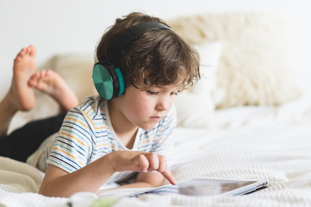 Chico lindo acostado en la cama en casa y jugando con una tableta o leyendo en línea y escuchando música con auriculares inalámbricos