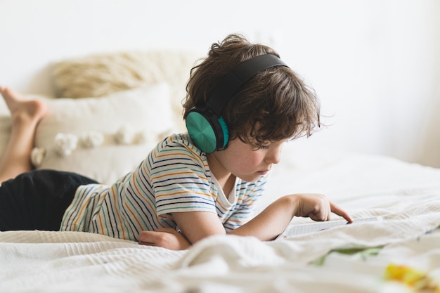 Chico lindo acostado en la cama en casa y jugando con una tableta o leyendo en línea y escuchando música con auriculares inalámbricos