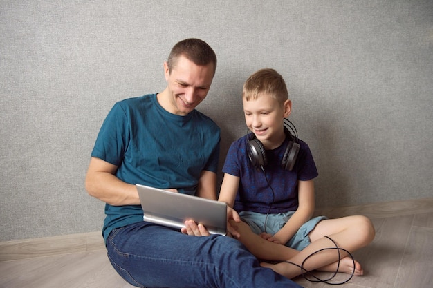 Un chico lindo con auriculares está sentado en el suelo con su padre y mira la tableta sonriendo