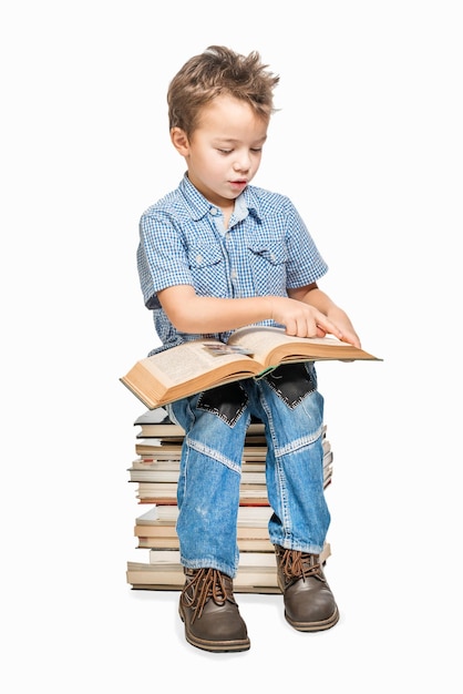Chico lindo con una camisa azul sentado en una pila de libros y leyendo un libro sobre un fondo blanco aislado