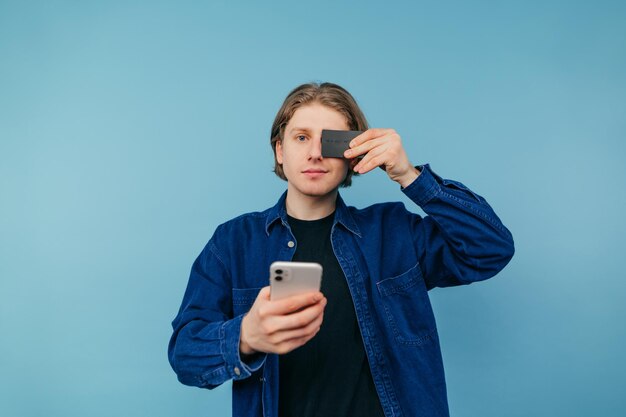 Chico lindo en una camisa con un teléfono inteligente y una tarjeta bancaria en sus manos se encuentra sobre un fondo azul.