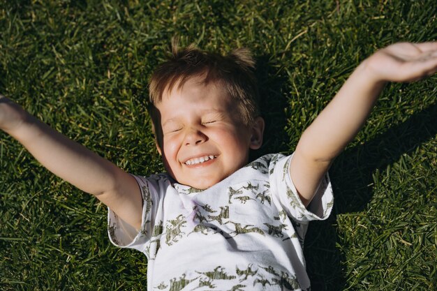 Foto chico lindo en camiseta blanca tendido sobre la hierba verde con las manos en alto y entrecerrar los ojos al sol