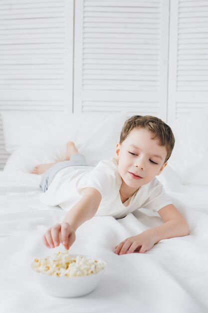 Chico lindo comiendo palomitas de maíz sentado en la cama con sábanas blancas