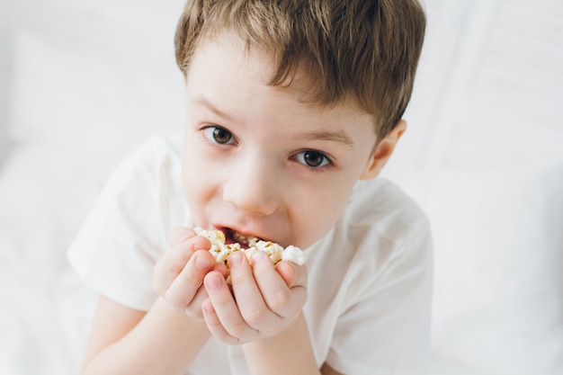 Chico lindo comiendo palomitas de maíz sentado en la cama con sábanas blancas