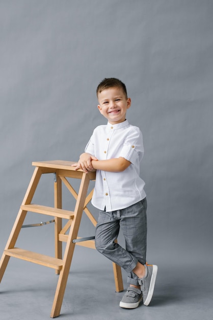 Un chico lindo y elegante se apoya en una escalera de tijera de madera sobre un fondo gris y sonríe