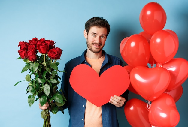 Chico lindo esperando novia con regalos de San Valentín, sosteniendo un ramo de rosas y corazón rojo, sonriendo a la cámara, de pie sobre fondo azul.